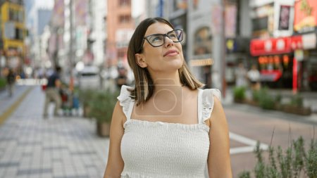 Photo for Globe-trotting beauty, stunning adult hispanic woman with glasses, looking around the vibrant tokyo cityscape. this brunette tourist is mesmerized by japan's urban architecture. - Royalty Free Image