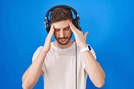 Photo for Hispanic man with beard listening to music wearing headphones with hand on head, headache because stress. suffering migraine. - Royalty Free Image