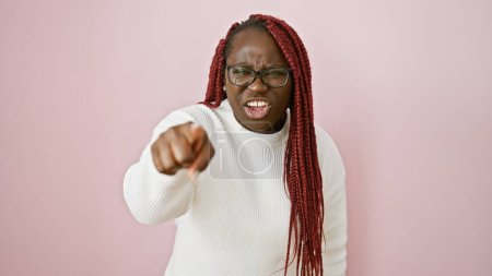 Mujer afroamericana enojada con trenzas apuntando con el dedo sobre un fondo rosa aislado