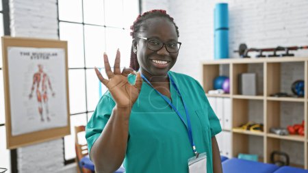 Photo for Smiling african american woman in scrubs gesturing okay in a physiotherapy clinic - Royalty Free Image