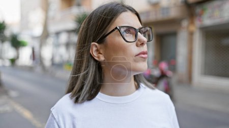Foto de Una joven contemplativa hispana con gafas se encuentra en una calle urbana con un fondo borroso de la ciudad - Imagen libre de derechos