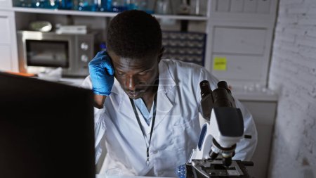 Photo for African male scientist feeling exhausted in the lab, leaning on microscope and computer - Royalty Free Image