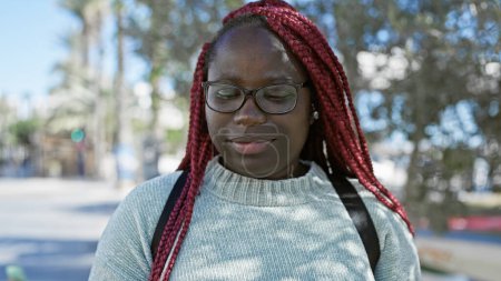 Photo for African american woman with braids wearing glasses stands thoughtfully on a sunny urban street. - Royalty Free Image