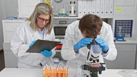 Photo for Man and woman, two concentrated scientists together in a lab, sitting, taking notes during microscope analysis - Royalty Free Image