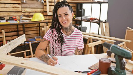 Photo for Confident, smiling hispanic woman, an amputee carpenter, drawing confidently with pen at her carpentry workshop - Royalty Free Image