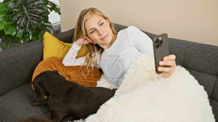 Téléchargez les photos : Femme blonde se détendre sur le canapé avec labrador noir dans le salon confortable, prendre selfie - en image libre de droit