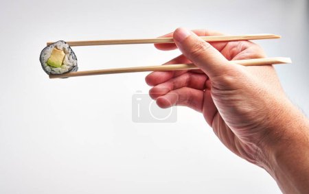 Photo for A man expertly grips a sushi roll with chopsticks against a white background, highlighting japanese cuisine and dexterity. - Royalty Free Image