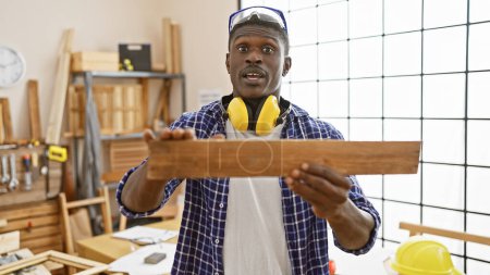 Foto de Carpintero afroamericano examina la madera en un taller bien iluminado con equipo de seguridad. - Imagen libre de derechos