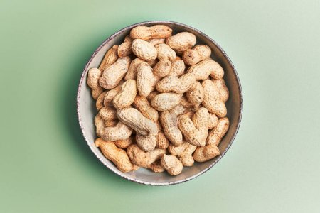 Overhead view of unshelled peanuts in a ceramic bowl on a green background, implying healthy snacking.