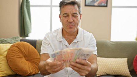 Foto de Hombre de mediana edad contando dólares de Singapur en una acogedora sala de estar, ilustrando las finanzas en casa. - Imagen libre de derechos