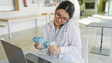 Téléchargez les photos : Une jeune femme hispanique examine la monnaie brésilienne dans un cadre de bureau moderne, reflétant le travail financier. - en image libre de droit
