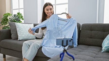 Photo for A cheerful young hispanic woman showcases a blue shirt to a smartphone on a tripod in a cozy, well-lit living room. - Royalty Free Image