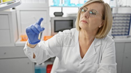 Foto de Mujer rubia madura inspecciona un vial en un entorno de laboratorio clínico. - Imagen libre de derechos