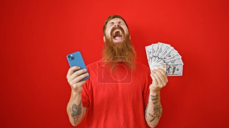 Cheerful young redhead guy beaming over his winnings, clutching dollars and smartphone against an isolated red backdrop!