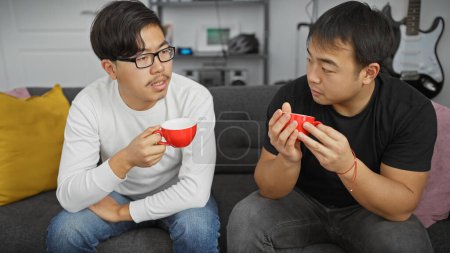 Téléchargez les photos : Deux hommes asiatiques ayant une conversation dans un salon moderne tout en tenant des tasses à café rouges - en image libre de droit