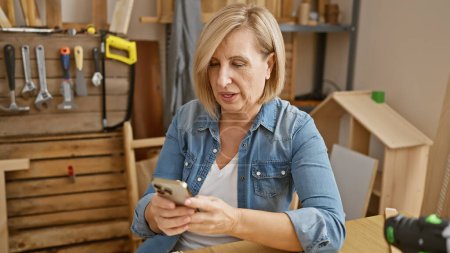 Photo for Mature caucasian woman using smartphone in a woodwork shop surrounded by tools and wooden furniture. - Royalty Free Image