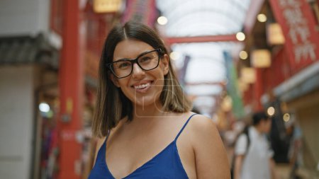 Belle jeune femme hispanique portant des lunettes debout souriant sur le marché japonais