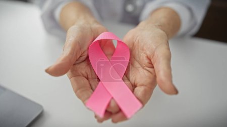 Reife Frau hält rosa Schleife in Klinik und symbolisiert Brustkrebs-Bewusstsein und Unterstützung.