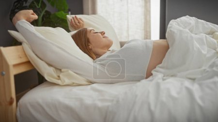 A peaceful young caucasian woman stretches while waking up in her cozy bedroom, embodying relaxation.