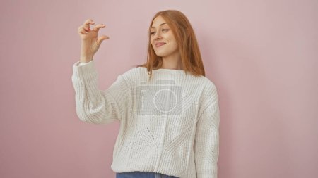 Smiling redhead woman in white sweater gesturing tiny amount over pink background