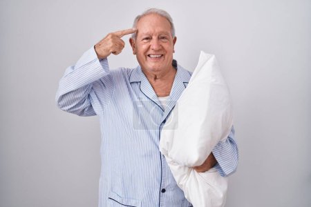 Foto de Hombre mayor con el pelo gris usando pijama abrazando almohada sonriendo apuntando a la cabeza con un dedo, gran idea o pensamiento, buena memoria - Imagen libre de derechos