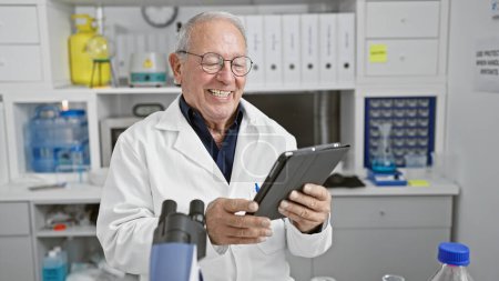 Téléchargez les photos : Homme scientifique senior souriant confiant en utilisant le pavé tactile au laboratoire - en image libre de droit