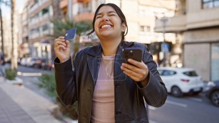 Foto de Una alegre joven hispana con tarjeta de crédito y teléfono inteligente en una animada calle de la ciudad. - Imagen libre de derechos