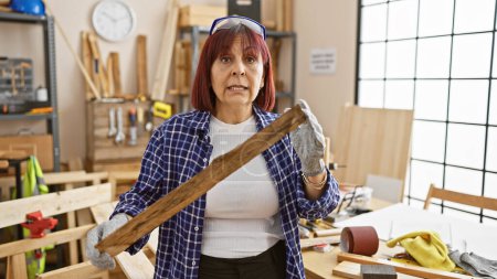 Photo for Middle-aged hispanic woman working in a carpentry workshop, holding a wooden plank. - Royalty Free Image