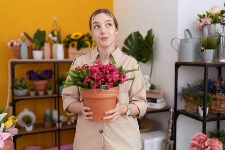 Téléchargez les photos : Jeune femme caucasienne travaillant au magasin de fleuriste tenant pot de plante souriant regardant vers le côté et regardant loin en pensant. - en image libre de droit
