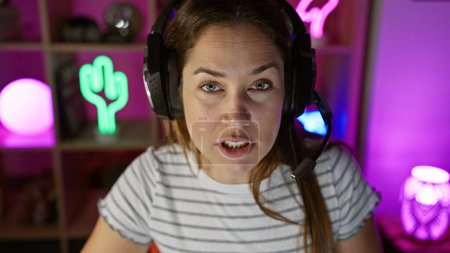 A focused woman with headphones in a colorful gaming room looks intently at the screen.