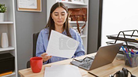 Photo for A professional young woman reviews documents at her office desk with a laptop and accessories in the background. - Royalty Free Image