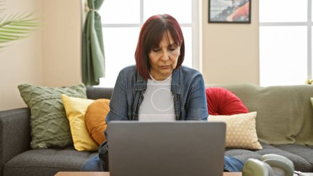 Foto de Mujer hispana mayor enfocada usando laptop en una acogedora sala de estar adornada con cojines coloridos. - Imagen libre de derechos