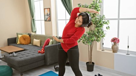 Foto de Una mujer en forma se ejercita en interiores, mostrando una sala de estar moderna mientras usa auriculares y ropa deportiva. - Imagen libre de derechos