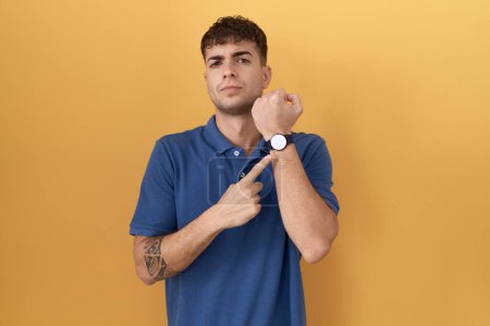 Photo for Young hispanic man standing over yellow background in hurry pointing to watch time, impatience, looking at the camera with relaxed expression - Royalty Free Image