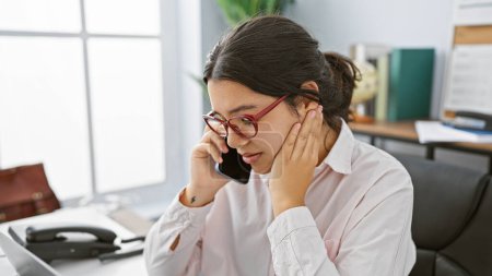 Foto de Una joven hispana profesional en gafas usando un teléfono en un entorno de oficina moderno. - Imagen libre de derechos