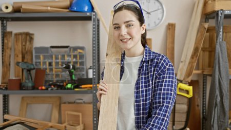 Foto de Mujer sonriente en taller sosteniendo madera con herramientas en el fondo. - Imagen libre de derechos