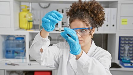 Foto de Una mujer hispana enfocada con cabello rizado analiza un tubo de ensayo en un entorno de laboratorio, destacando la importancia de la investigación científica. - Imagen libre de derechos