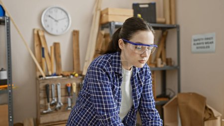 Photo for Focused young woman wearing safety glasses in a carpentry workshop surrounded by tools and wood. - Royalty Free Image