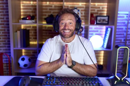Middle age man with beard playing video games wearing headphones praying with hands together asking for forgiveness smiling confident. 