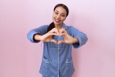 Téléchargez les photos : Jeune femme arabe portant un pyjama bleu souriant en amour faisant forme de symbole de coeur avec les mains. concept romantique. - en image libre de droit