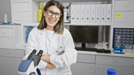 Téléchargez les photos : Jeune femme souriante avec des lunettes debout en toute confiance dans un laboratoire, portant un blouse de laboratoire avec les bras croisés. - en image libre de droit