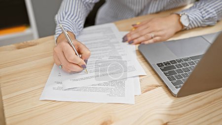 Photo for A professional woman signs a document in a modern office environment, showcasing a laptop and striped shirt. - Royalty Free Image