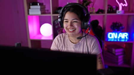 A smiling young hispanic woman wearing headphones in a neon-lit gaming room at night.