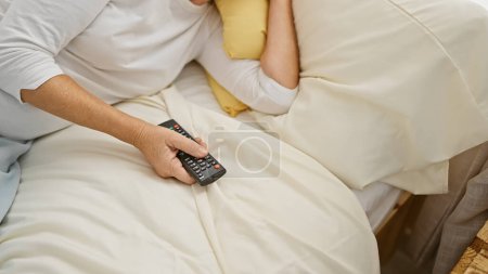 Téléchargez les photos : Image recadrée d'une femme d'âge moyen tenant une télécommande alors qu'elle était couchée au lit à la maison - en image libre de droit