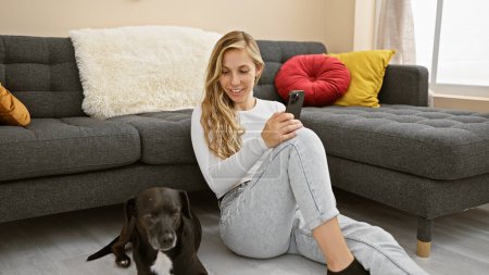 Téléchargez les photos : Une jeune femme souriante utilisant un smartphone à côté d'un labrador noir détendu dans un salon confortable. - en image libre de droit