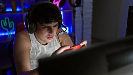 Téléchargez les photos : Jeune homme concentré portant un casque jouant à des jeux vidéo dans une salle de jeux sombre la nuit - en image libre de droit