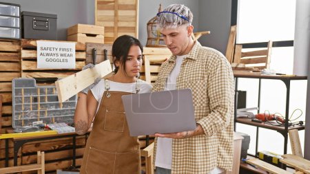 Man and woman in a carpentry workshop reviewing a project on a laptop amidst woodworking tools and workbenches.