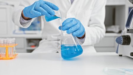 Foto de Un científico masculino en un laboratorio vertiendo líquido azul de un vaso de precipitados en un frasco. - Imagen libre de derechos