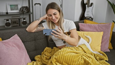 Téléchargez les photos : Femme souriante se détend sur un canapé avec son chien tout en utilisant un smartphone à la maison. - en image libre de droit