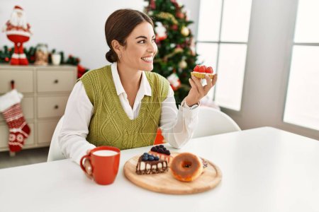 Young beautiful hispanic woman having breakfast sitting by christmas tree at home